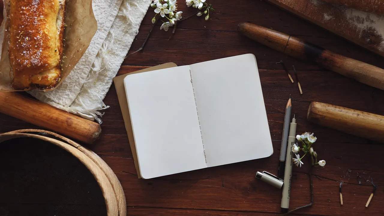 Carnet ouvert à une page blanche sur un plan de travail sombre, stylos posés à côté.