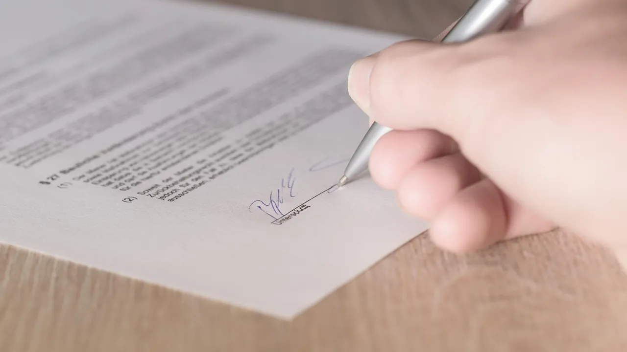 Main signant un contrat imprimé posé sur un plan de bois clair.